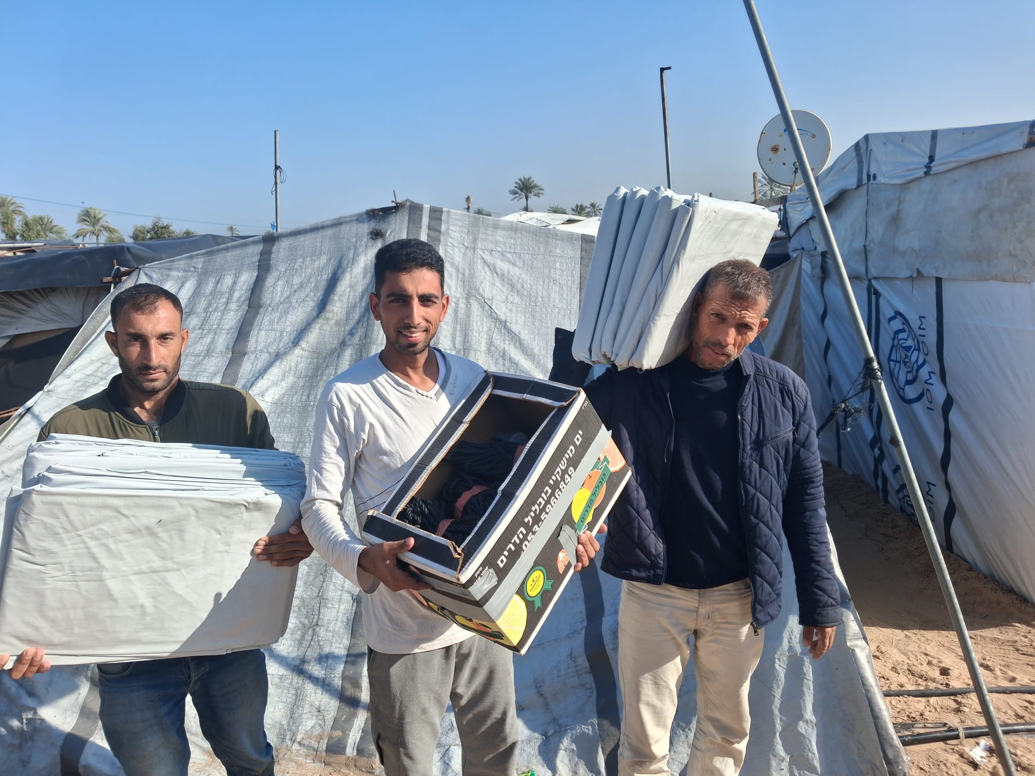 People carrying folded materials and supplies beside tents in Al Anwar