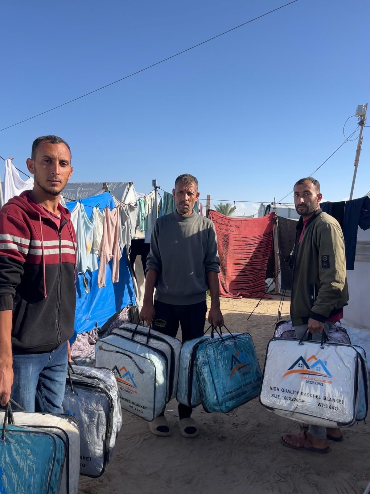 Three men holding packaged winter blankets beside tents at Al Anwar